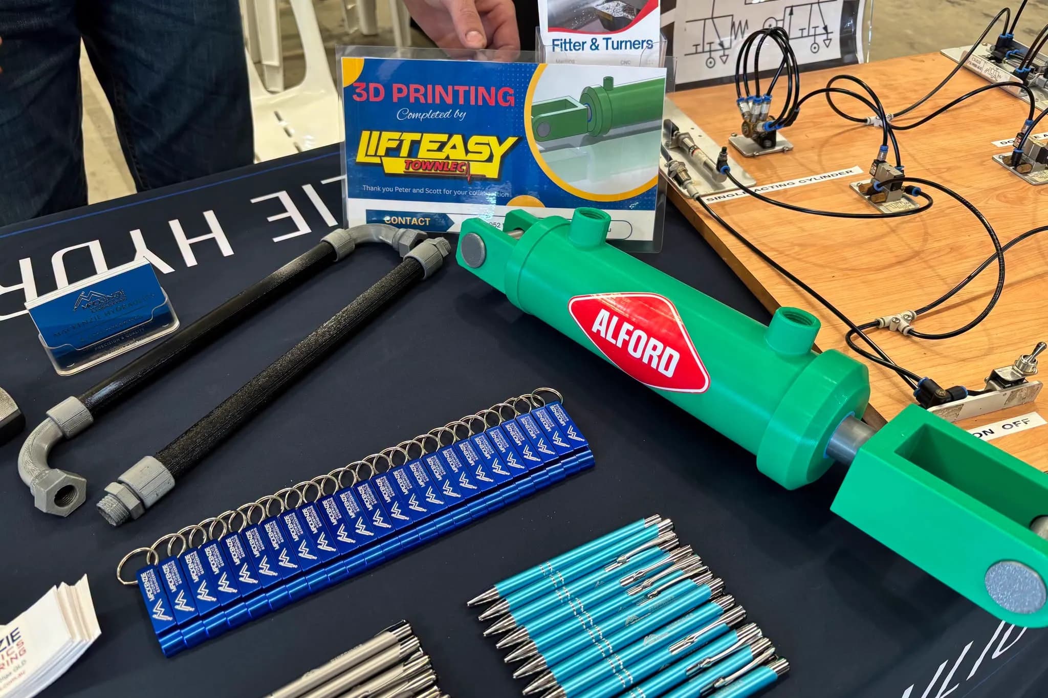 A 3D printed hydraulic cylinder model in green with printed hose fittings and elbows, laid out on the Mackenzie Hydraulics trade stand beside a card crediting the print