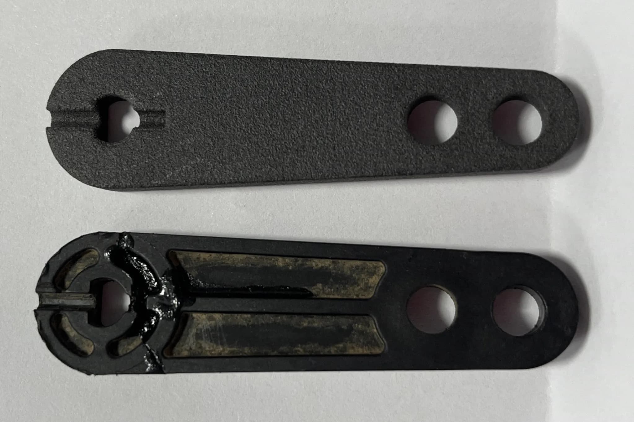 A matt black 3D printed part and the smooth black original it was copied from, the original split at its slotted end and holding old adhesive residue in two recesses, both with the same slot and two holes