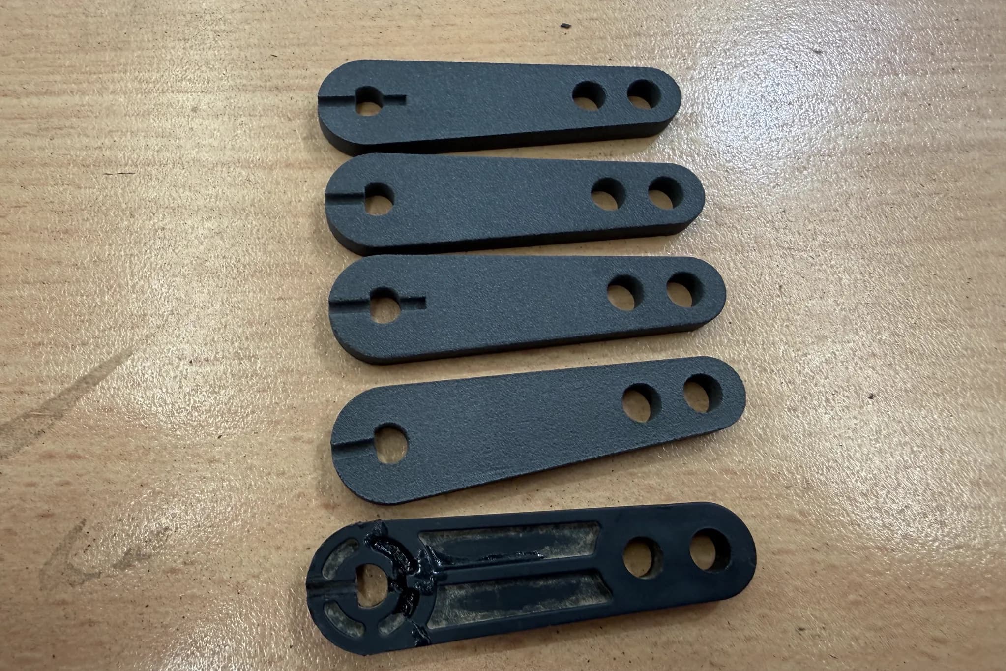 Four matt black 3D printed copies of a flat plastic part laid out on a Townlec workbench beside the black original they were drawn from