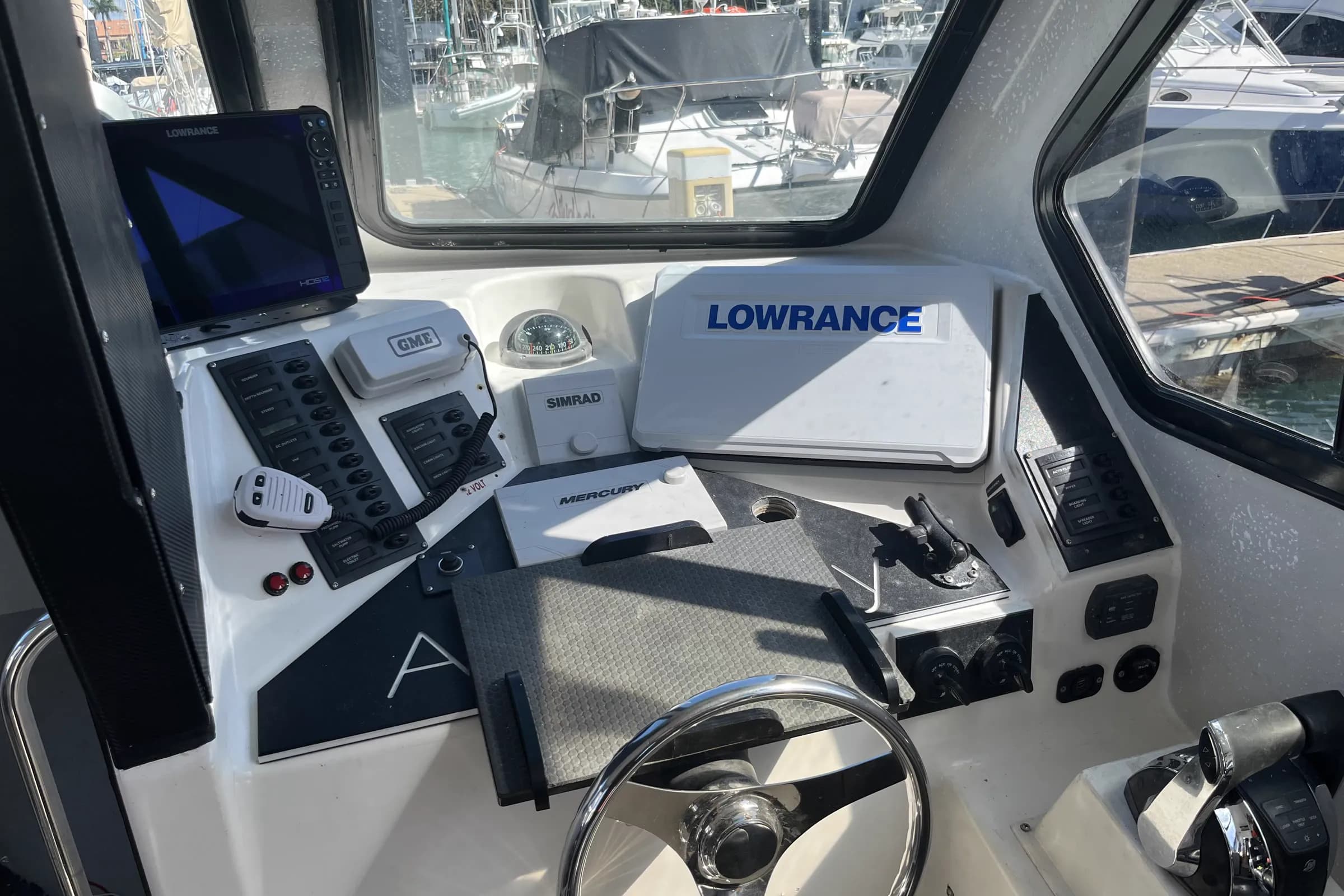 A boat helm at a marina with a Lowrance HDS12, a second Lowrance under its cover, a GME VHF, a Simrad autopilot control, a compass, Mercury controls and two labelled breaker panels