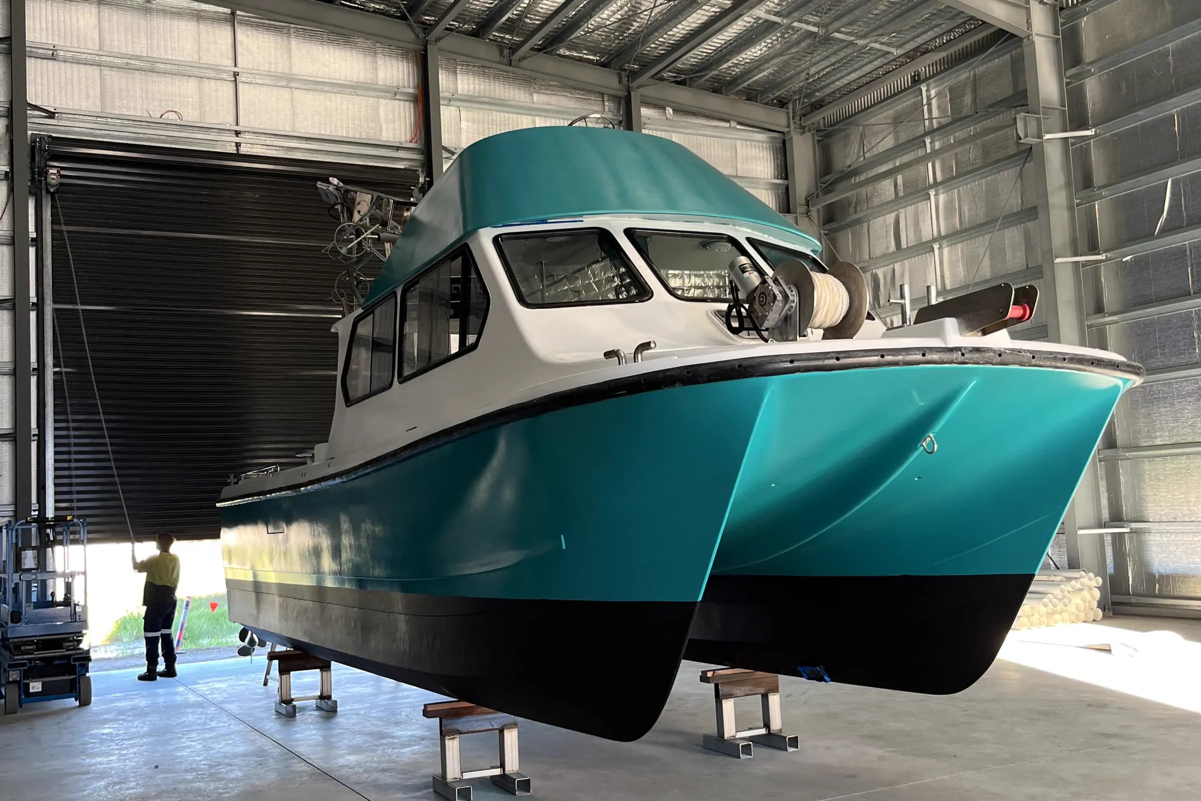 A teal and white cabin boat up on stands in a shed with its hull antifouled, seen from the bow