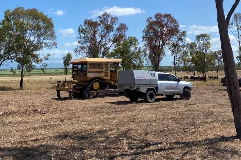 The silver Toyota LandCruiser 200 Series service ute parked beside a yellow tracked dozer in a dry North Queensland paddock, with cattle and ranges behind