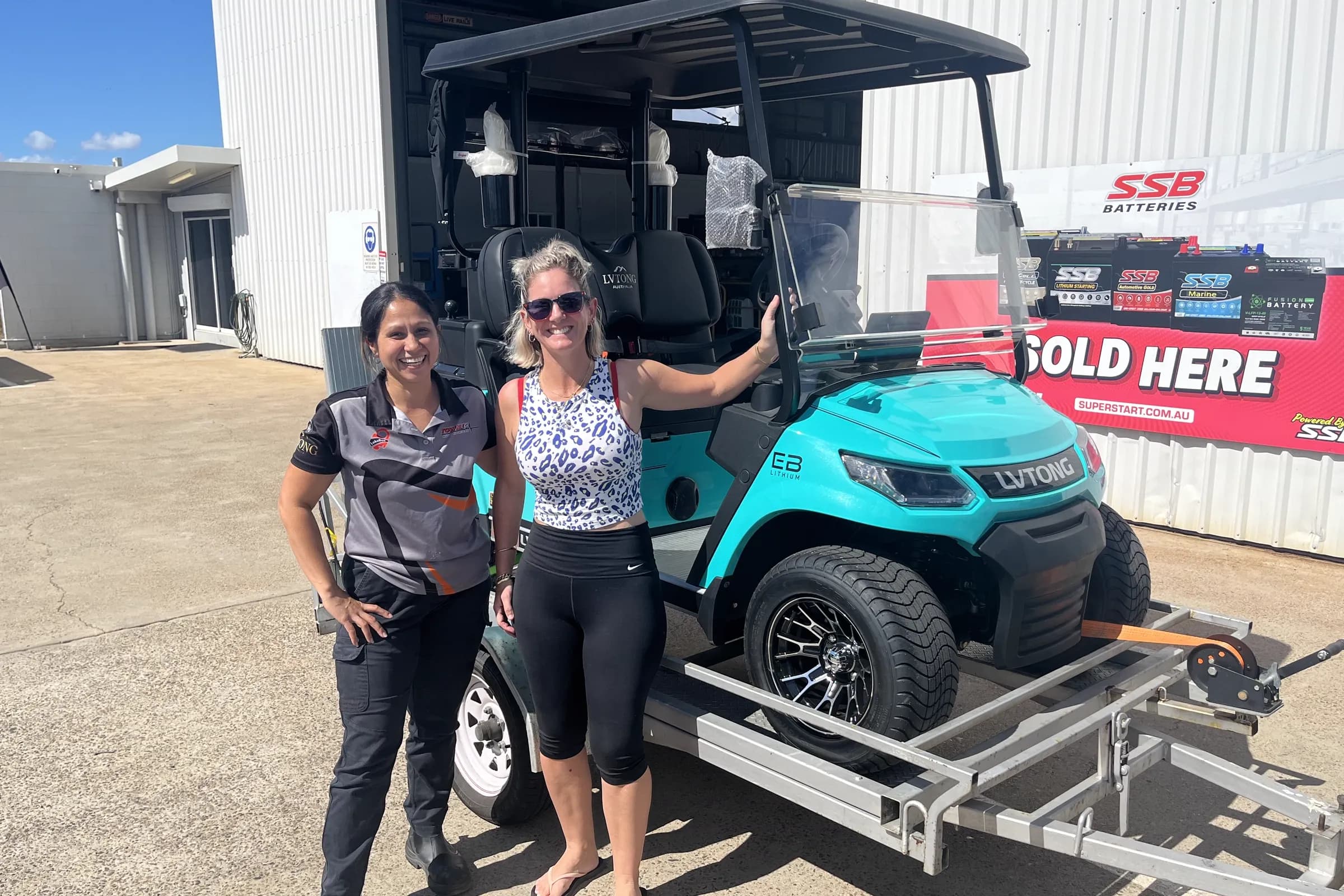 A Townlec staff member and a customer beside a teal LVTong golf cart strapped to a trailer outside the Townsville workshop, SSB battery banner behind