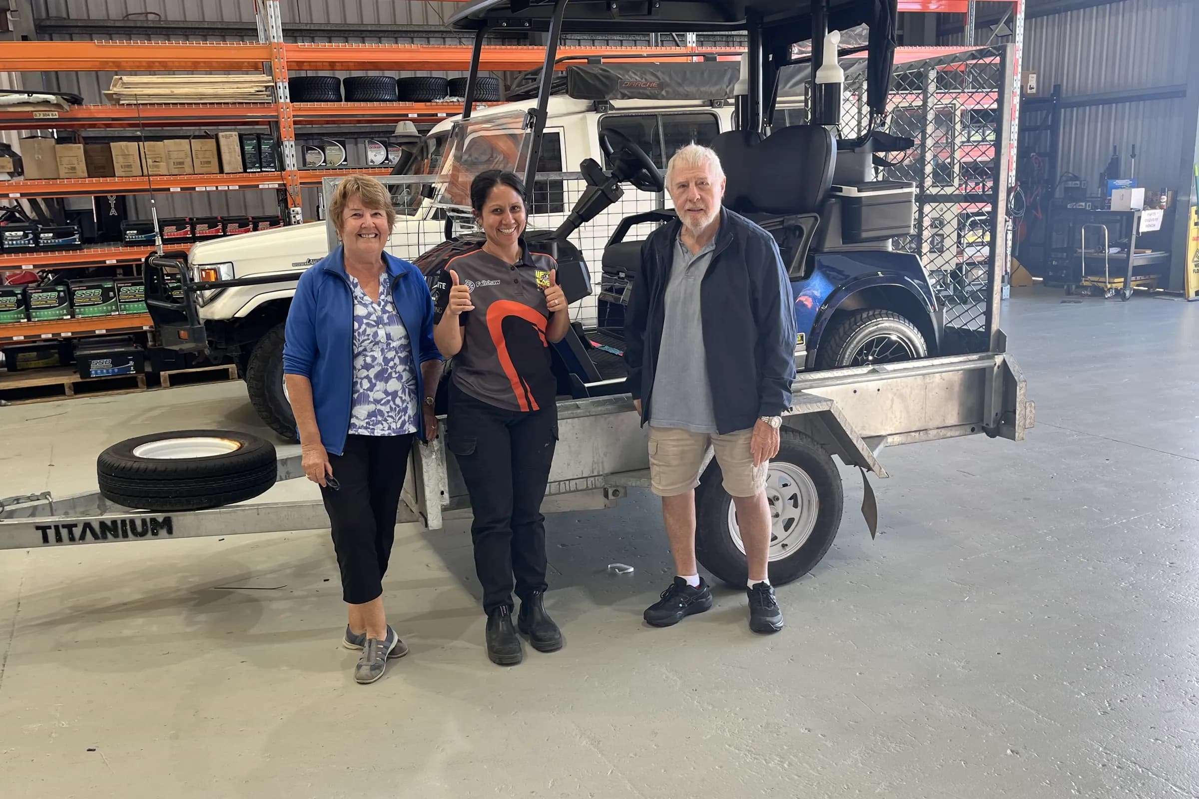 Two customers and a Townlec staff member beside a lifted dark blue golf cart with a cargo cage, loaded on a trailer inside the Townsville workshop