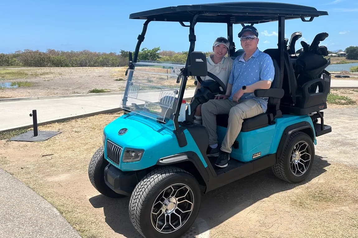 A couple sitting in a teal lithium golf cart on a North Queensland golf course, golf bags loaded on the rear rack and water beyond