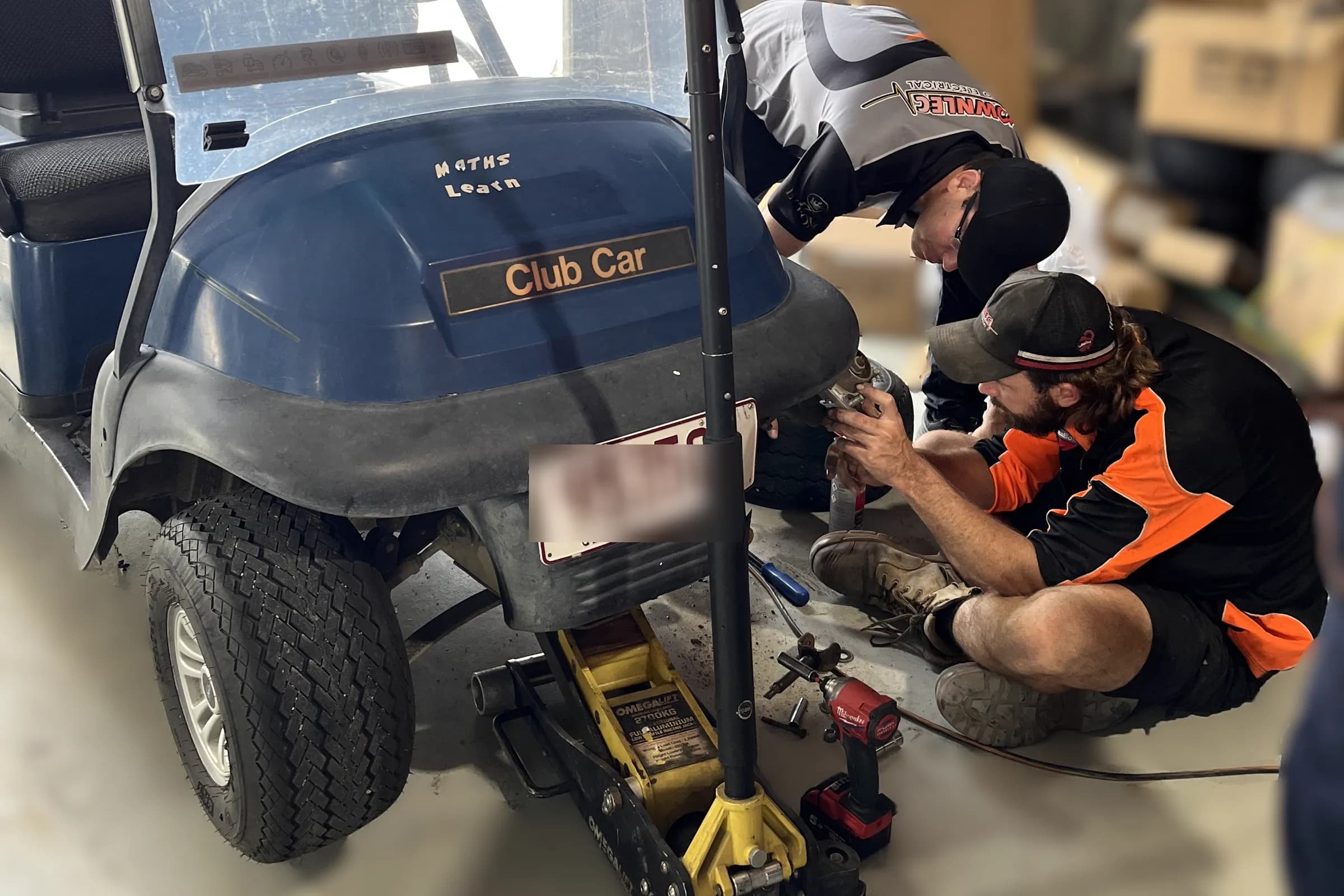 Two Townlec technicians working on a blue Club Car golf cart raised on a trolley jack in the Townsville workshop