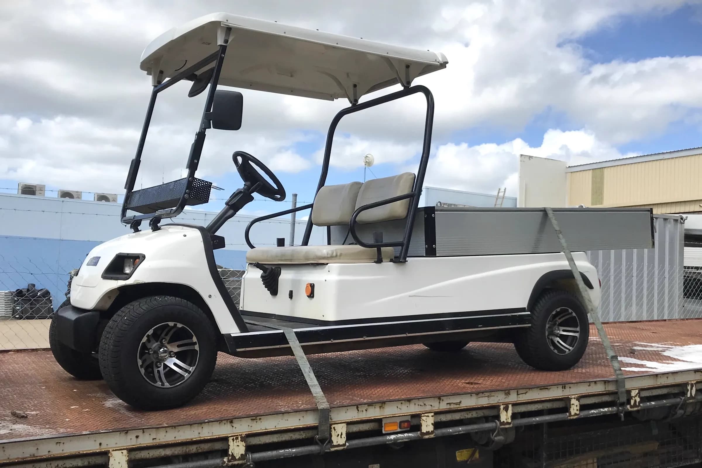 A used white utility golf cart with an alloy tray, strapped down on the back of a flatbed truck